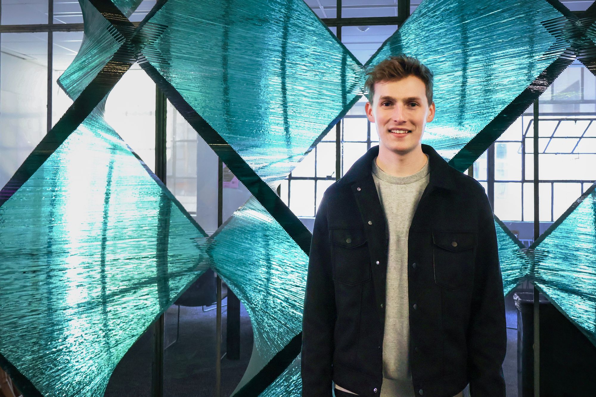 Young man standing and smiling in front of a large sculptural installation made of layered, translucent turquoise glass arranged in intersecting diamond-shaped patterns. The setting appears to be a modern indoor space with glass walls and natural light.
